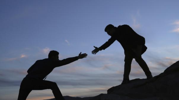 The silhouette of two people on a steep hike. The figure on the higher ground is reaching their hand out to help the figure on the lower ground climb.