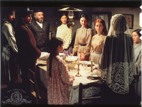 A scene from the movie Fiddler on the Roof, of a Jewish family celebrating the Sabbath meal. A woman wears a prayer shawl and lights candles, and her smiling family stands around the table.