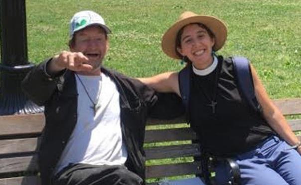 A man and a woman sit next to each other on a bench, both smiling. The man is homeless, wearing a T-shirt and a baseball cap, and the woman is a pastor wearing a clerical shirt and a wide brimmed straw hat.